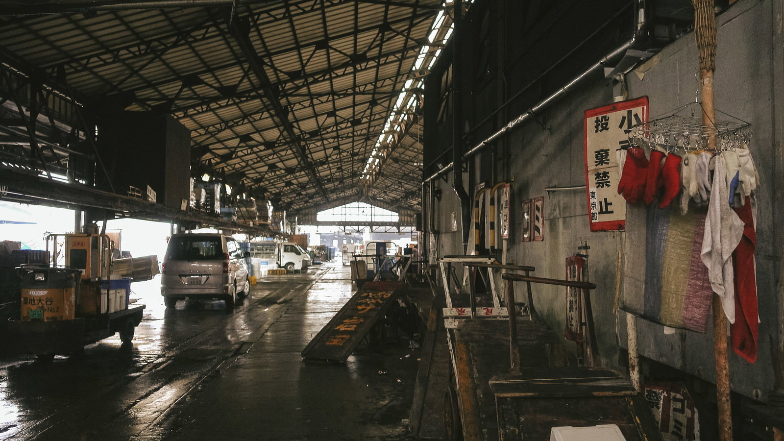 鉄骨屋根とコンクリート壁に囲まれた屋内作業場。車両と作業道具、干されたタオルが見える湿った床