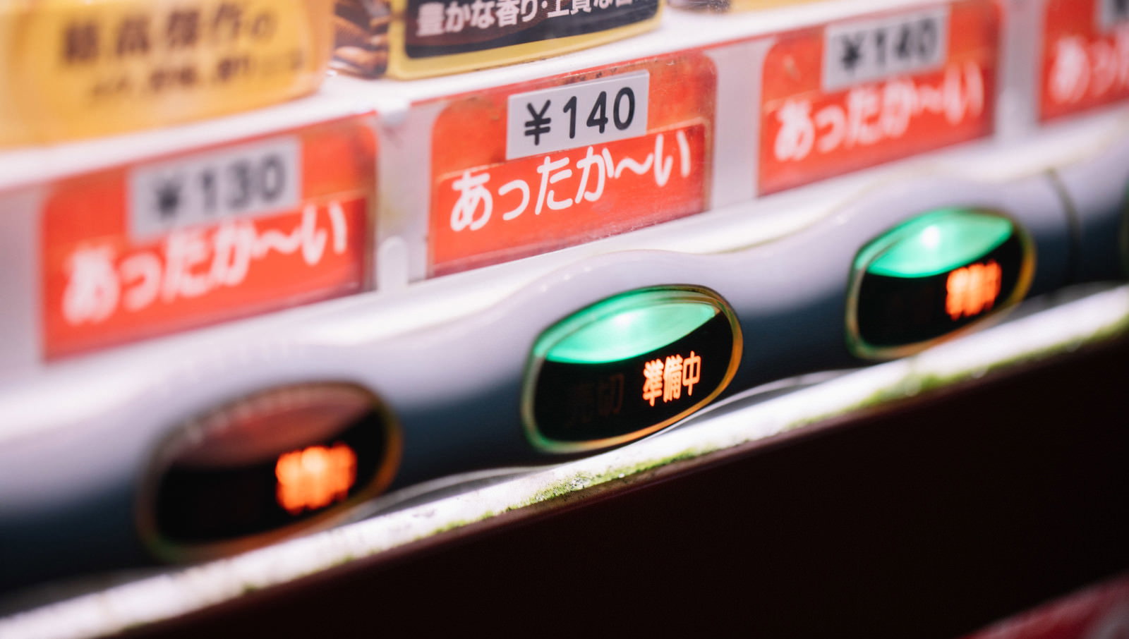 冬季に「あったか～い」のポップを貼った自動販売機。緑と赤のボタンが点灯し、価格表示が見える斜め前からの撮影