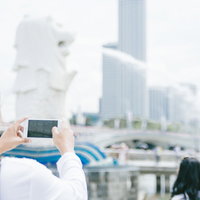 マーライオンの下で遠近法を使ってスマホで噴水を飲む姿を撮影する男性の写真