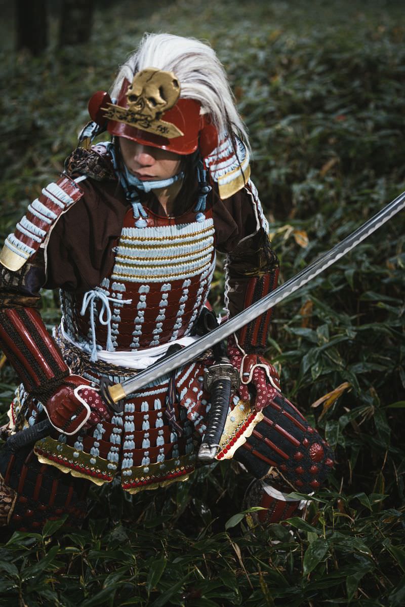 赤い甲冑姿で日本刀を構える白髪の侍が笹原に佇む［モデル：まーこ］