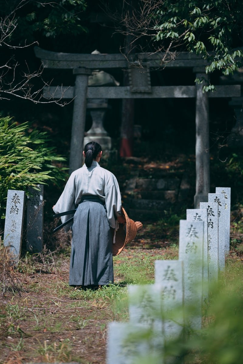 神社の鳥居前で袴を着た男性が石柱の間に立っている後ろ姿［モデル：まーこ］
