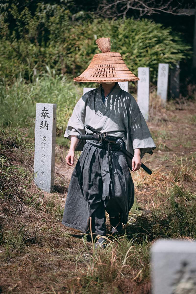 網代笠を被り袴姿の武士が奉納石柱の間を歩く様子［モデル：まーこ］