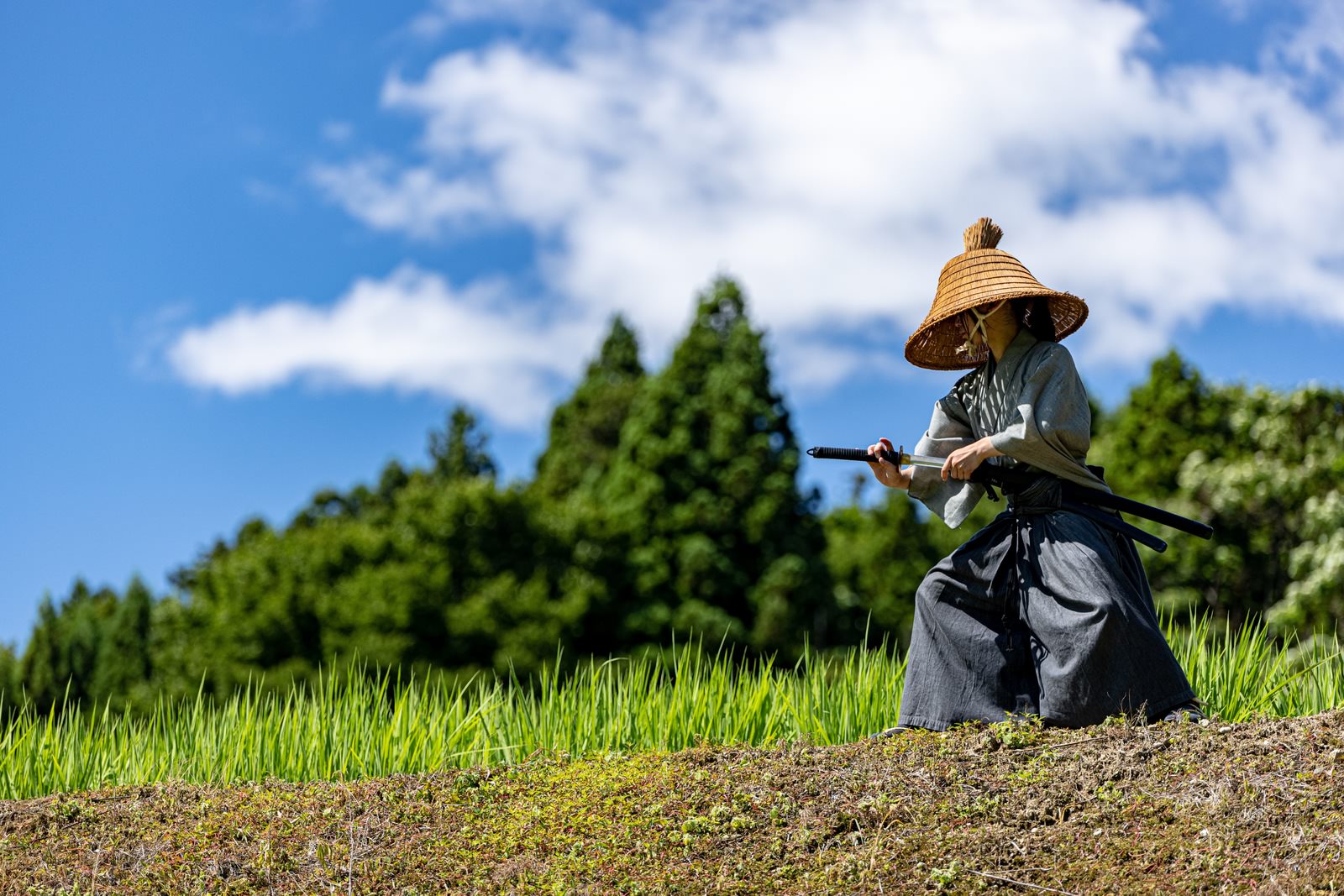 青空の下で網代笠をかぶった袴姿の侍が日本刀を抜刀している様子［モデル：まーこ］