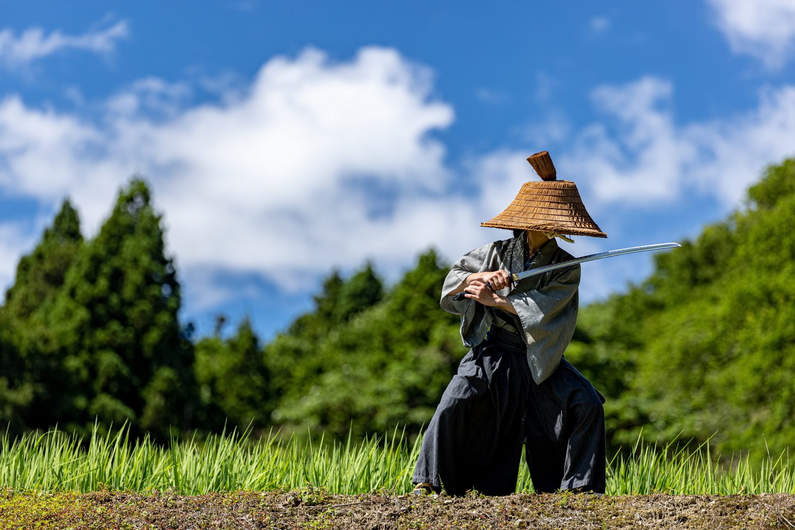 青空の下で日本刀を構える網代笠を被った黒袴姿の侍［モデル：まーこ］