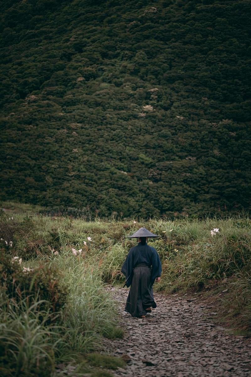A samurai wearing a conical hat descends a mountain path through the grass