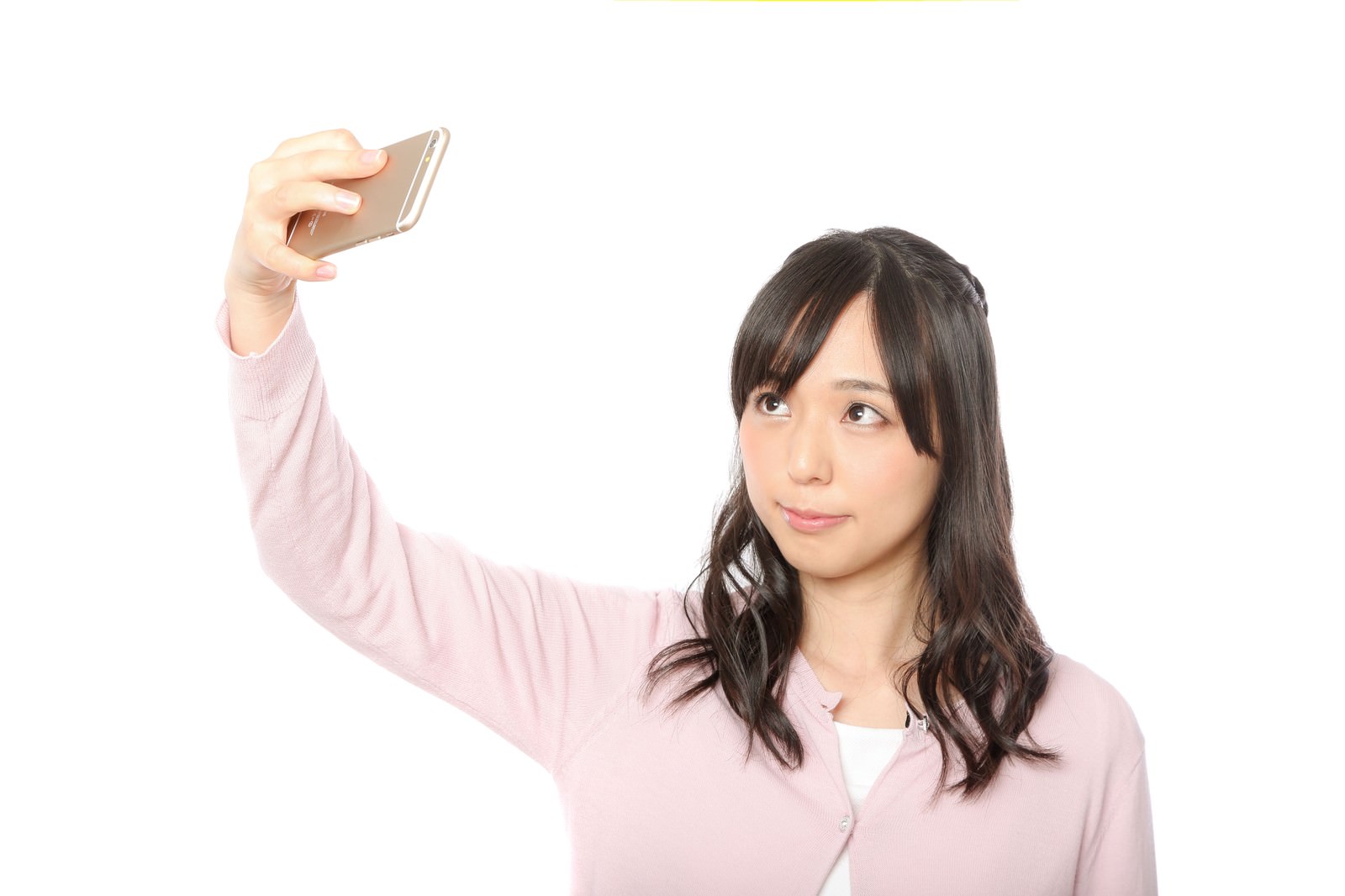 ピンク色のカーディガンを着た女性がスマートフォンを掲げて自撮りをしている［モデル：さとうゆい］