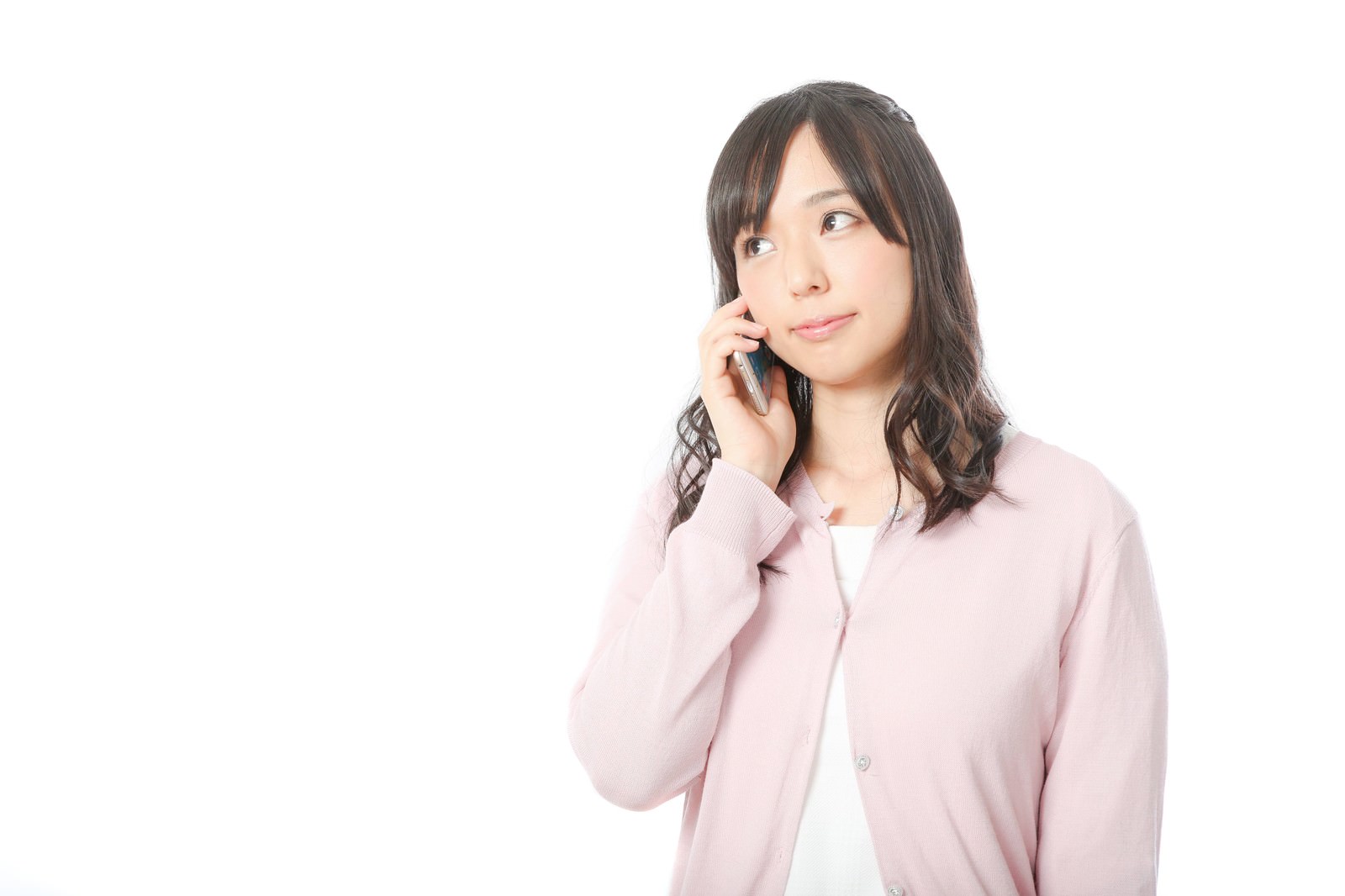 ピンクのカーディガンを着た女性がスマートフォンで電話をしながら笑顔で上を見つめている［モデル：さとうゆい］