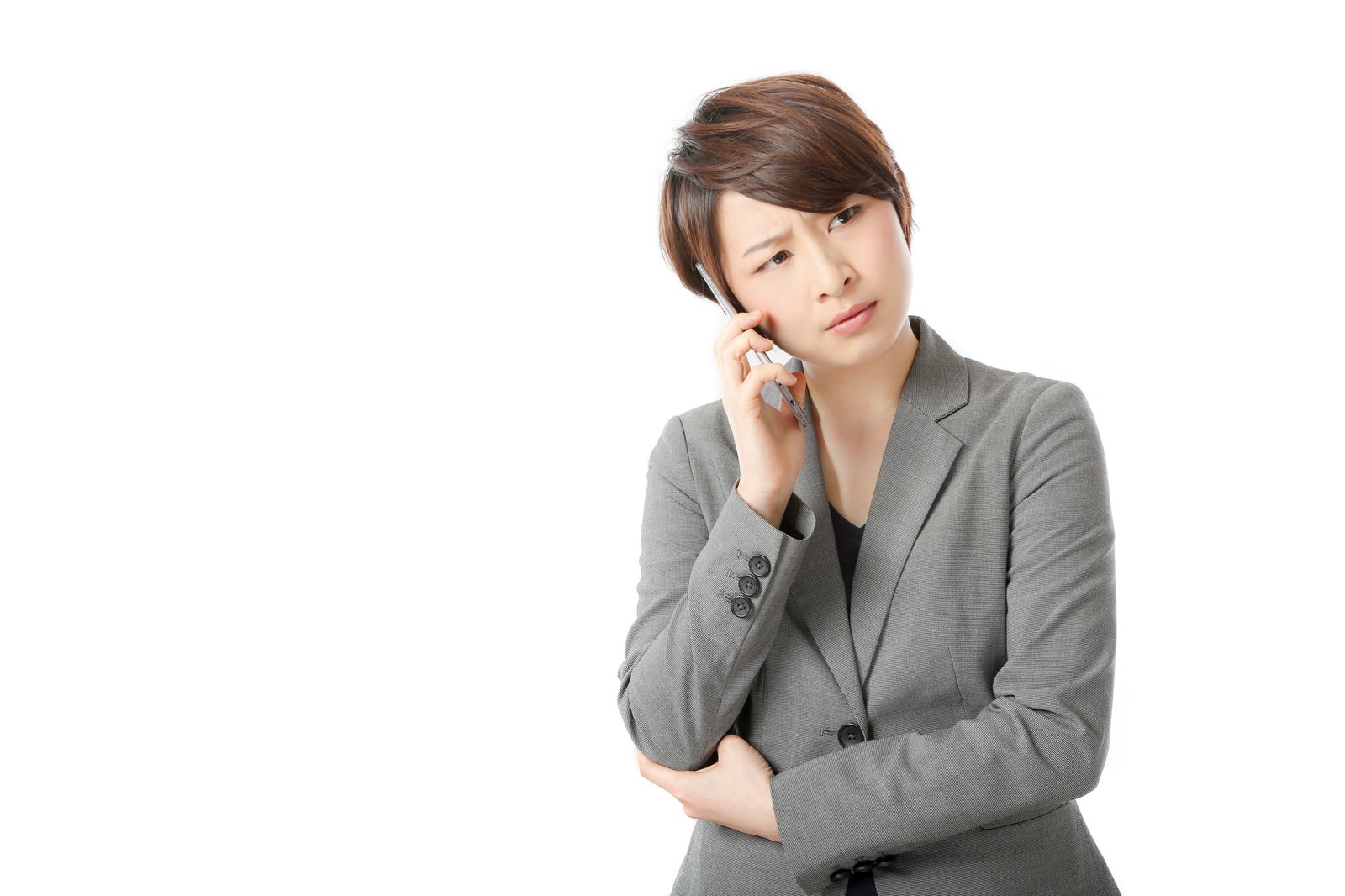 グレーのビジネスジャケットを着た女性がスマートフォンを耳に当てて通話しながら、眉をひそめて考え込んでいる［モデル：八木彩香］