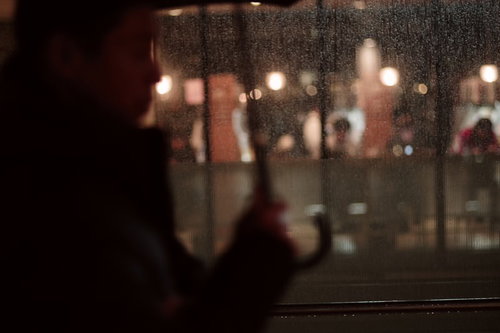傘をさす通行人が前を通りすぎる雨の夜