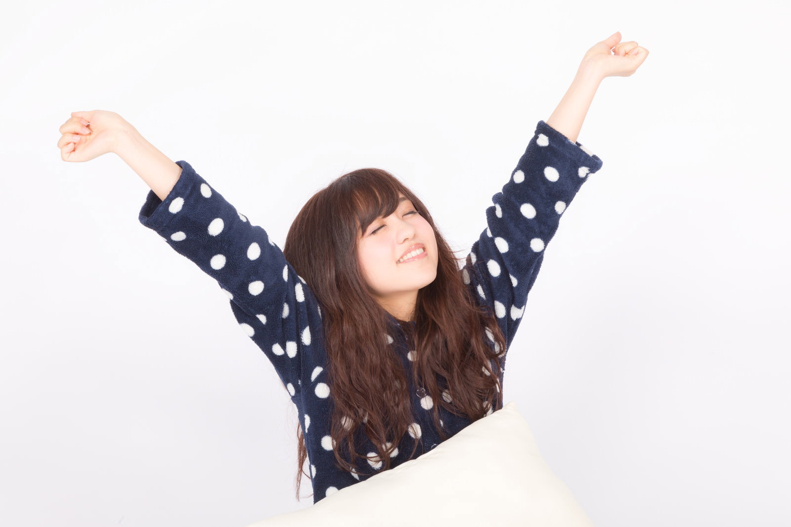 ドット柄のパジャマを着た女性が白い背景で両手を上げて伸びをしている［モデル：河村友歌］
