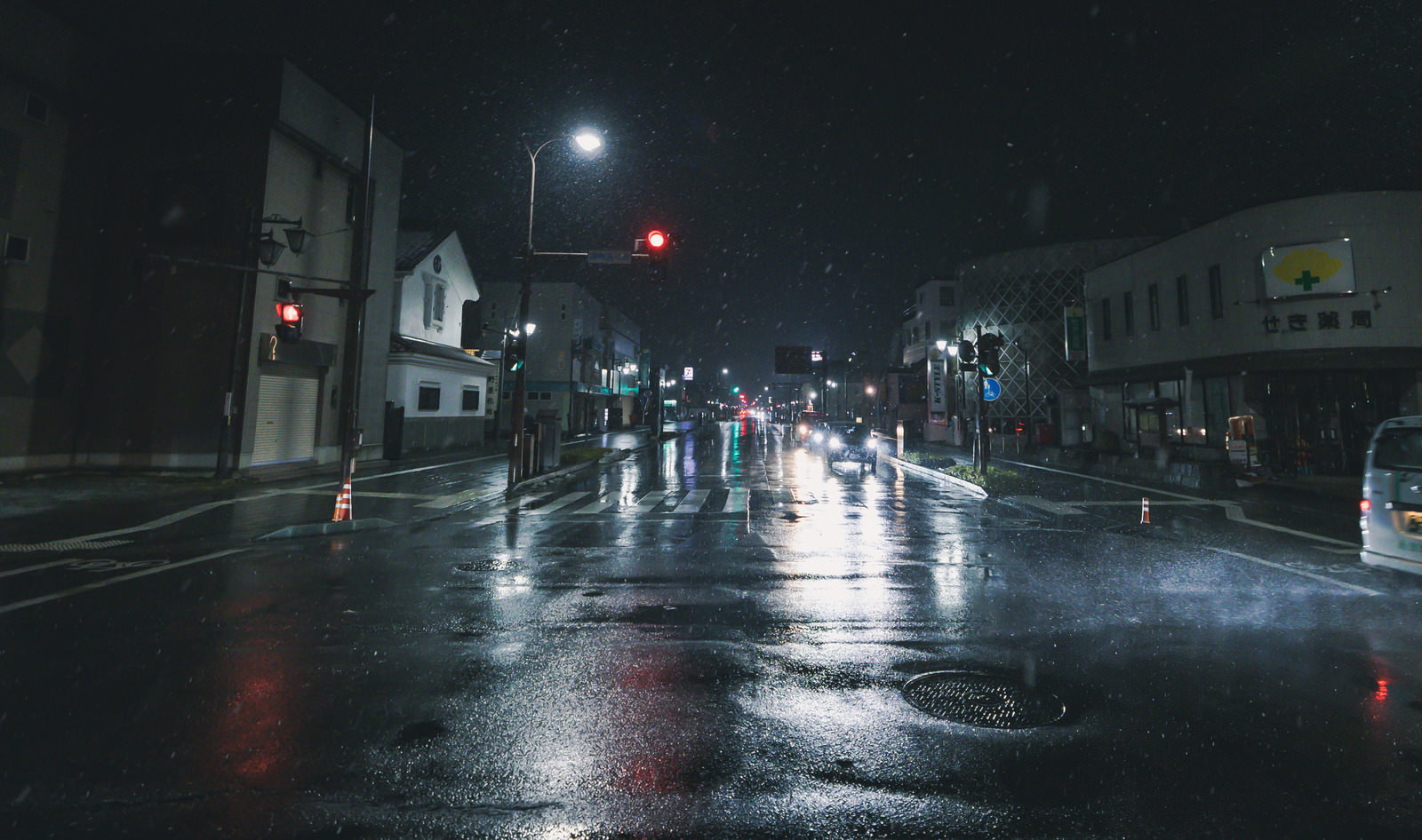 雨に濡れた深夜の交差点で、街灯と赤い信号機が照らす商店街の道路