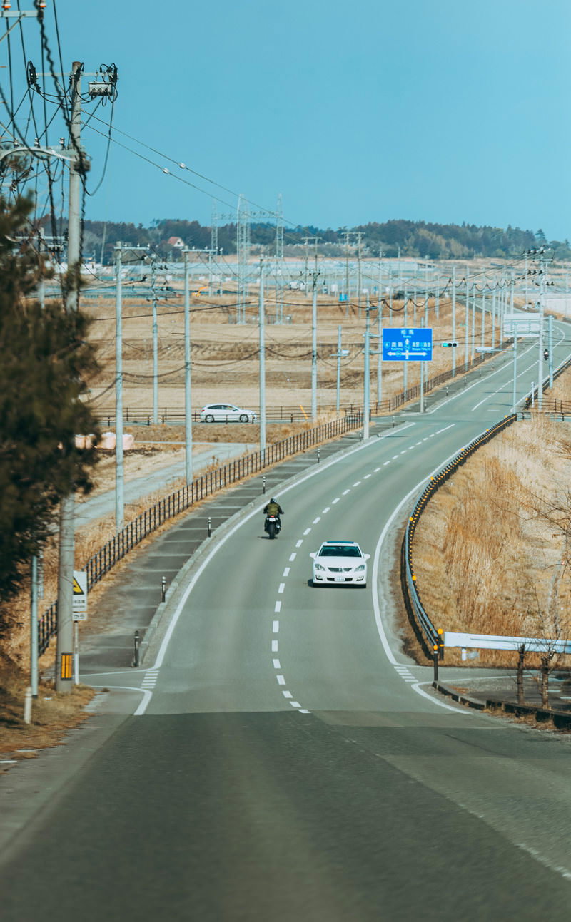 南相馬に向かう道路を走る白い乗用車とバイク。晴れた空と電柱のある風景。