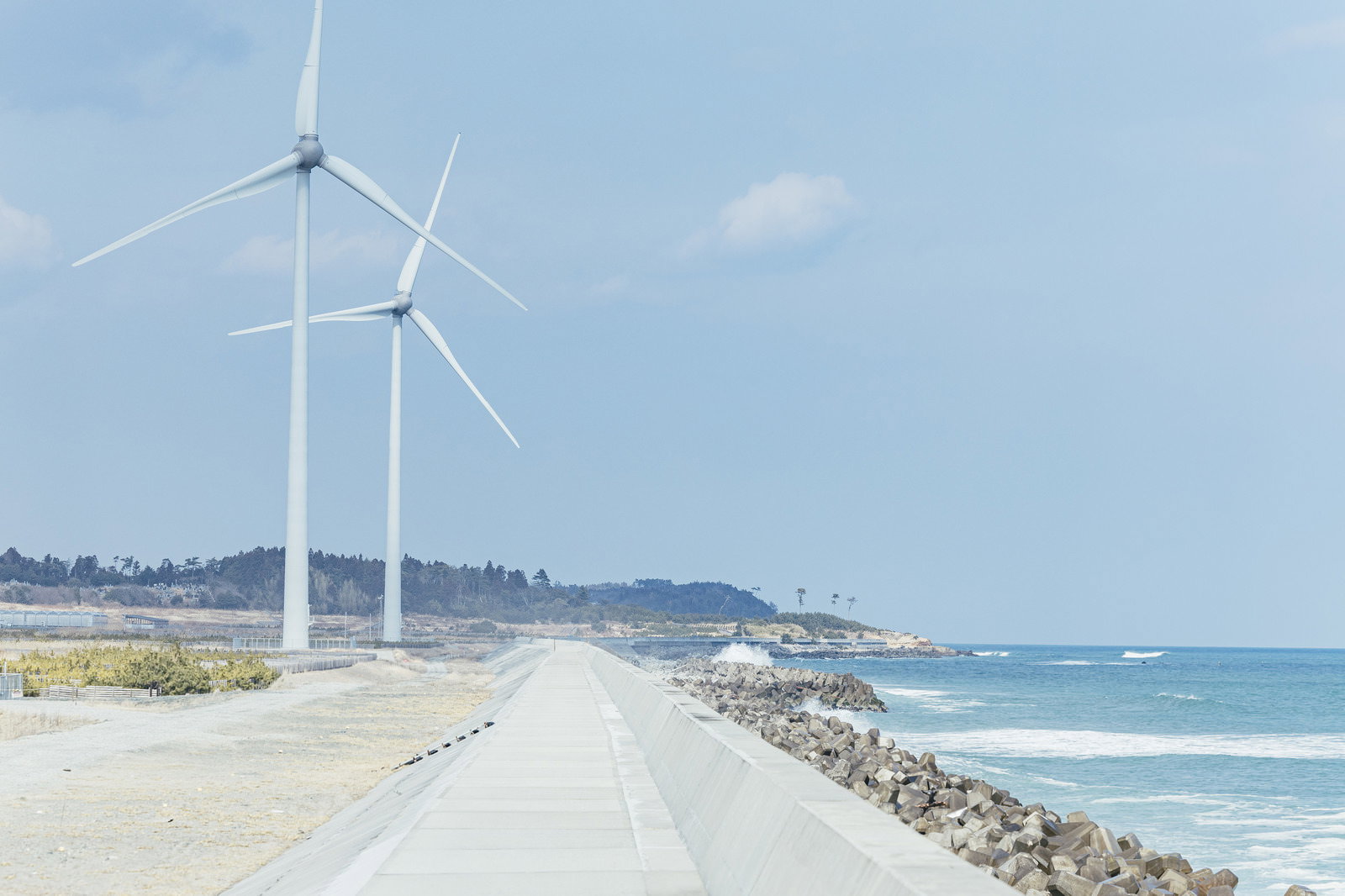 南相馬の海岸に立つ白い風力発電の風車と舗装路、防波堤、青い海