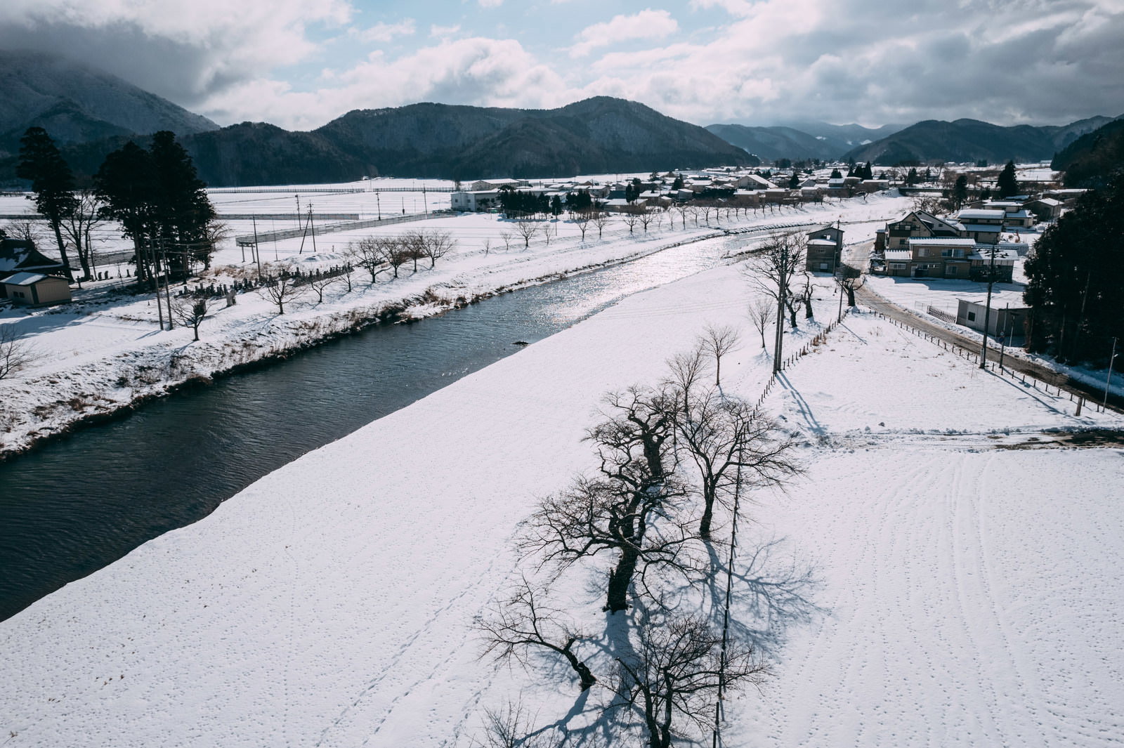 雪に覆われた舟津川と白い田畑、背景に雪をかぶった山々が見える郡山市の冬景色