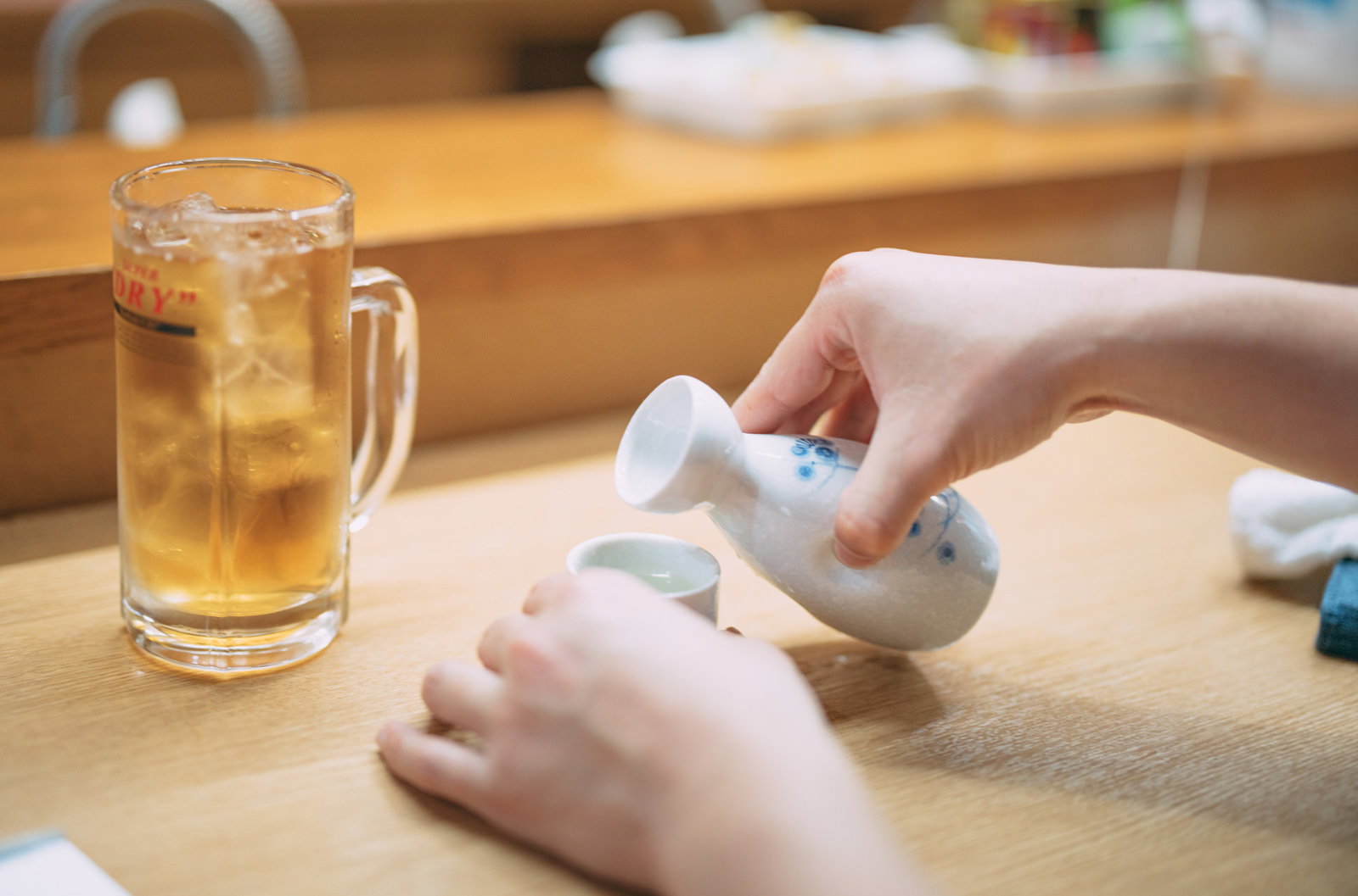 木製のカウンター上で、白いとっくりから琥珀色のお酒をグラスに注ぐ手