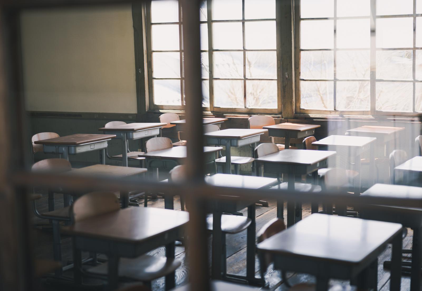 窓ガラス越しに見える複数の学習机と椅子が整列する教室の風景