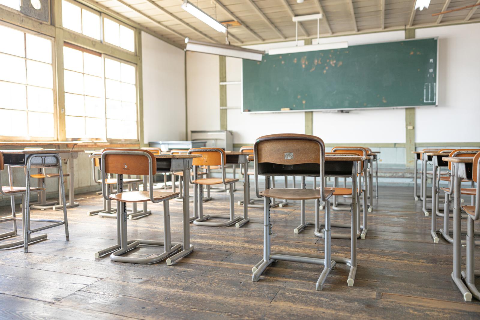 木製の学習机と椅子が並ぶ古い教室内に緑の黒板と教壇がある
