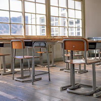 窓から差し込む光に照らされた学習机と椅子が並ぶ教室の写真