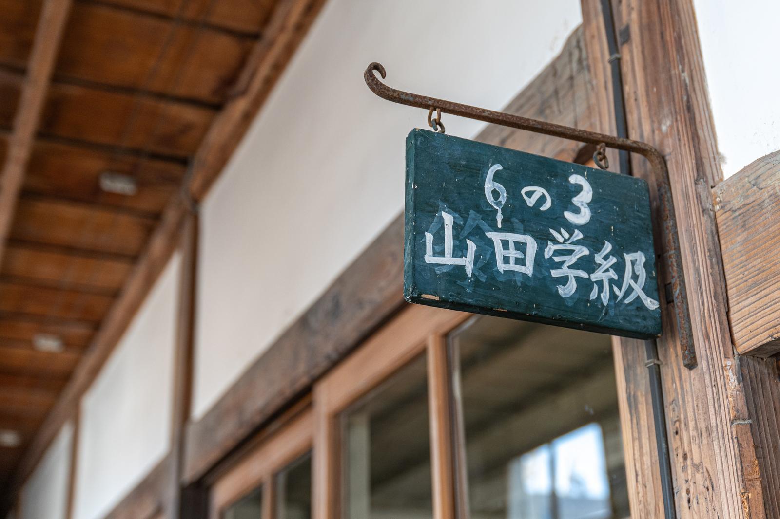 古い木造校舎の廊下に吊り下げられた緑の学級表示板に「6の3 山田学級」と白字で書かれている