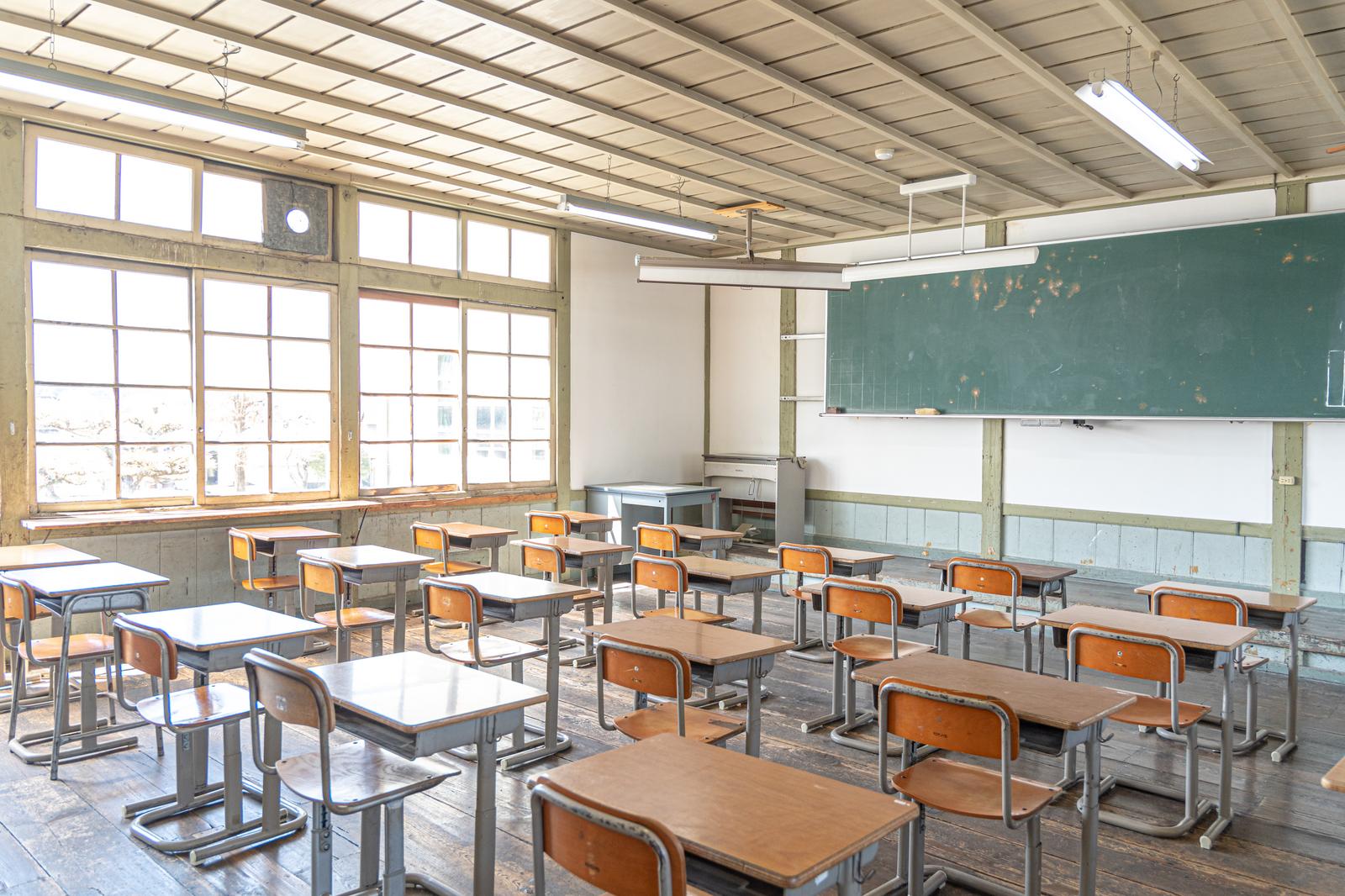 窓から光が入る古い教室。木製の学習椅子と机が並び、黒板が見える