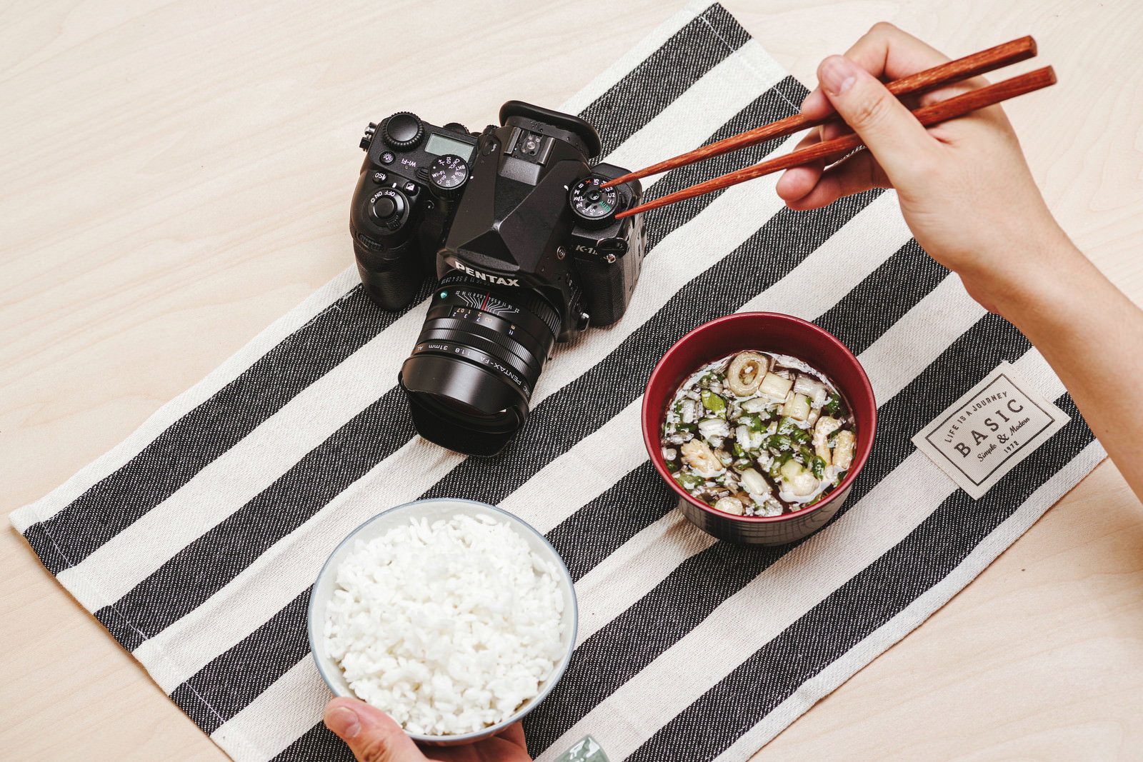 ストライプ柄のランチョンマットの上に置かれたPENTAXの一眼レフカメラと和食の朝食
