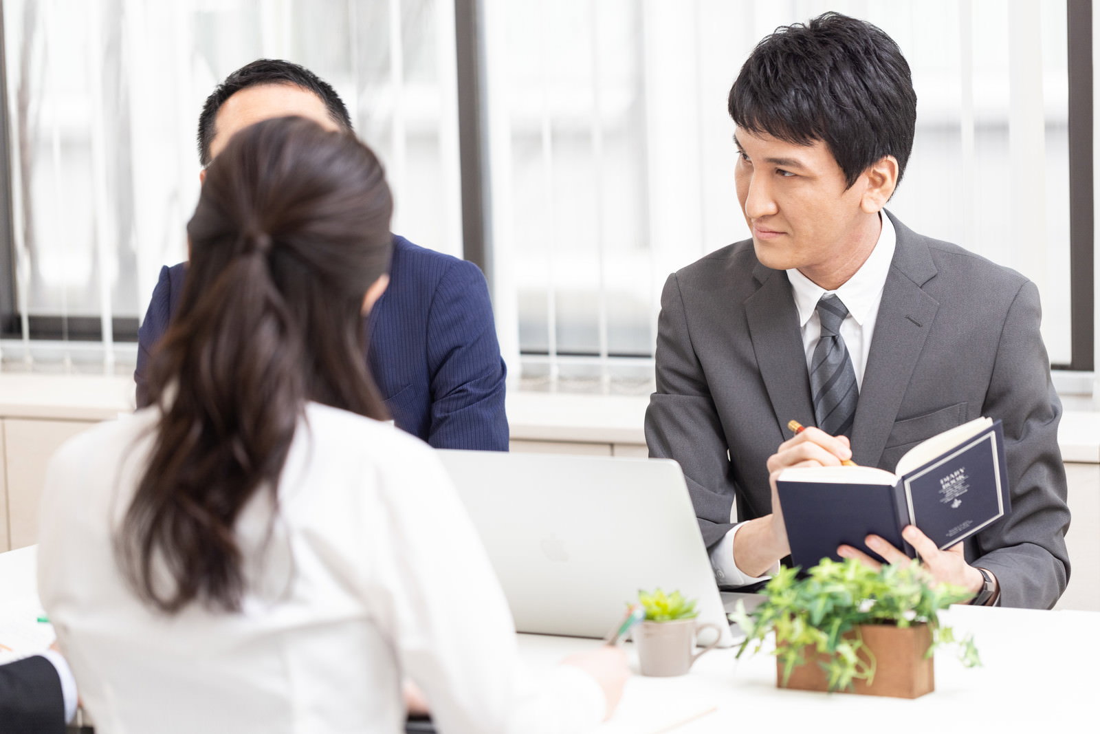 オフィスで会議をする男性が机に座り手帳や資料を見ながら話し合いをしている［モデル：ACE］