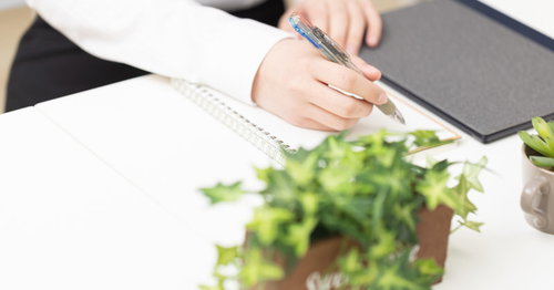 白いノートにペンでメモを書く女性の手元とデスク