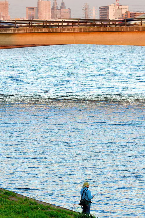 夕方の川岸で釣りをする釣り人と街並み