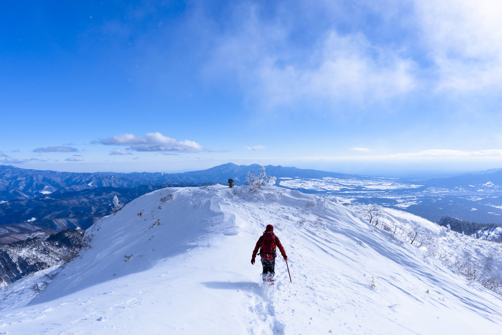 青空の下、雪に覆われた山稜を歩く赤いウェアの登山者と足跡