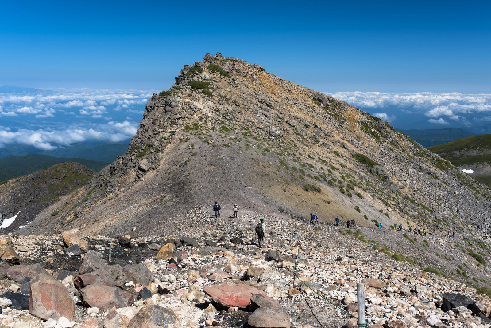 青空の下、乗鞍岳の岩場の登山道を山頂に向かって歩く登山者たち