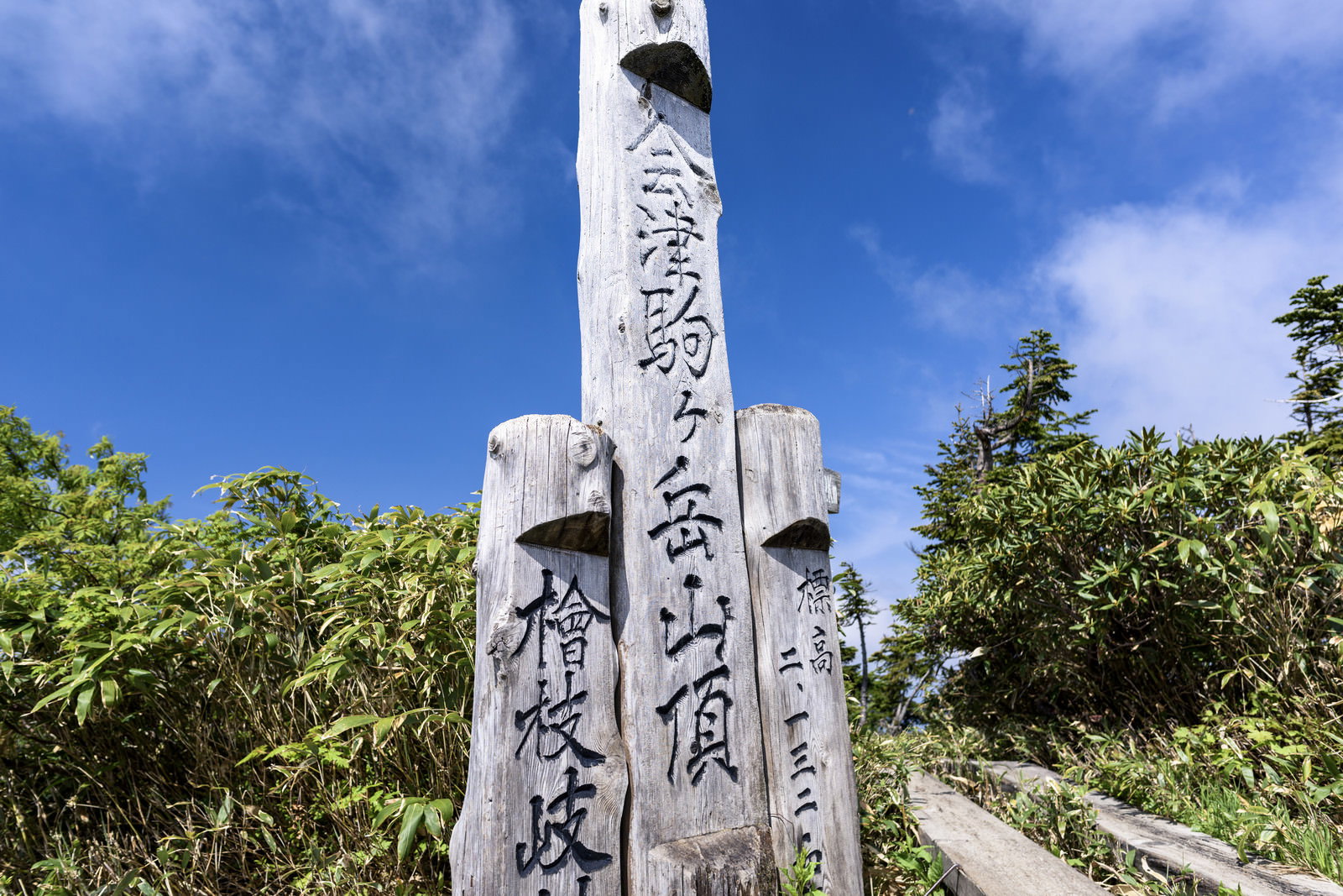 青空の下に立つ会津駒ヶ岳の白い山頂碑と周囲の緑豊かな山々