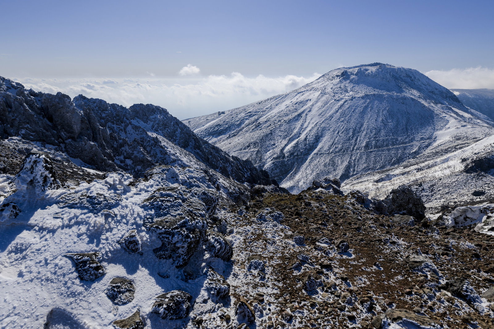 青空の下、雪化粧した那須茶臼岳と手前の雪が残る岩場の冬山風景