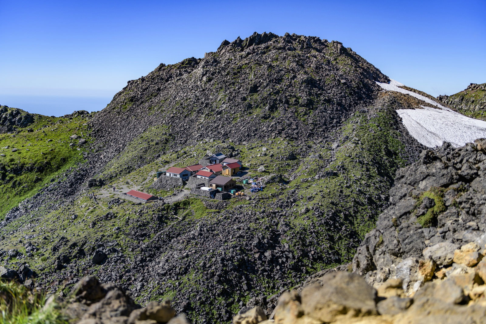 青空の下、岩場の斜面に建つ鳥海山の山小屋と周囲の緑豊かな高山植物