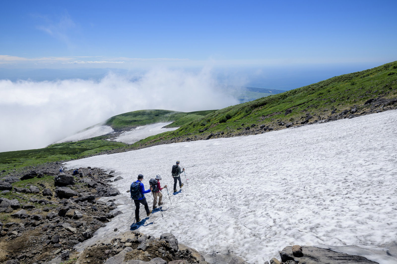 青空の下、鳥海山の雪渓を歩く3人の登山者