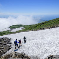 青空の下、鳥海山の雪渓を下る登山者たちの夏山トレッキングの写真