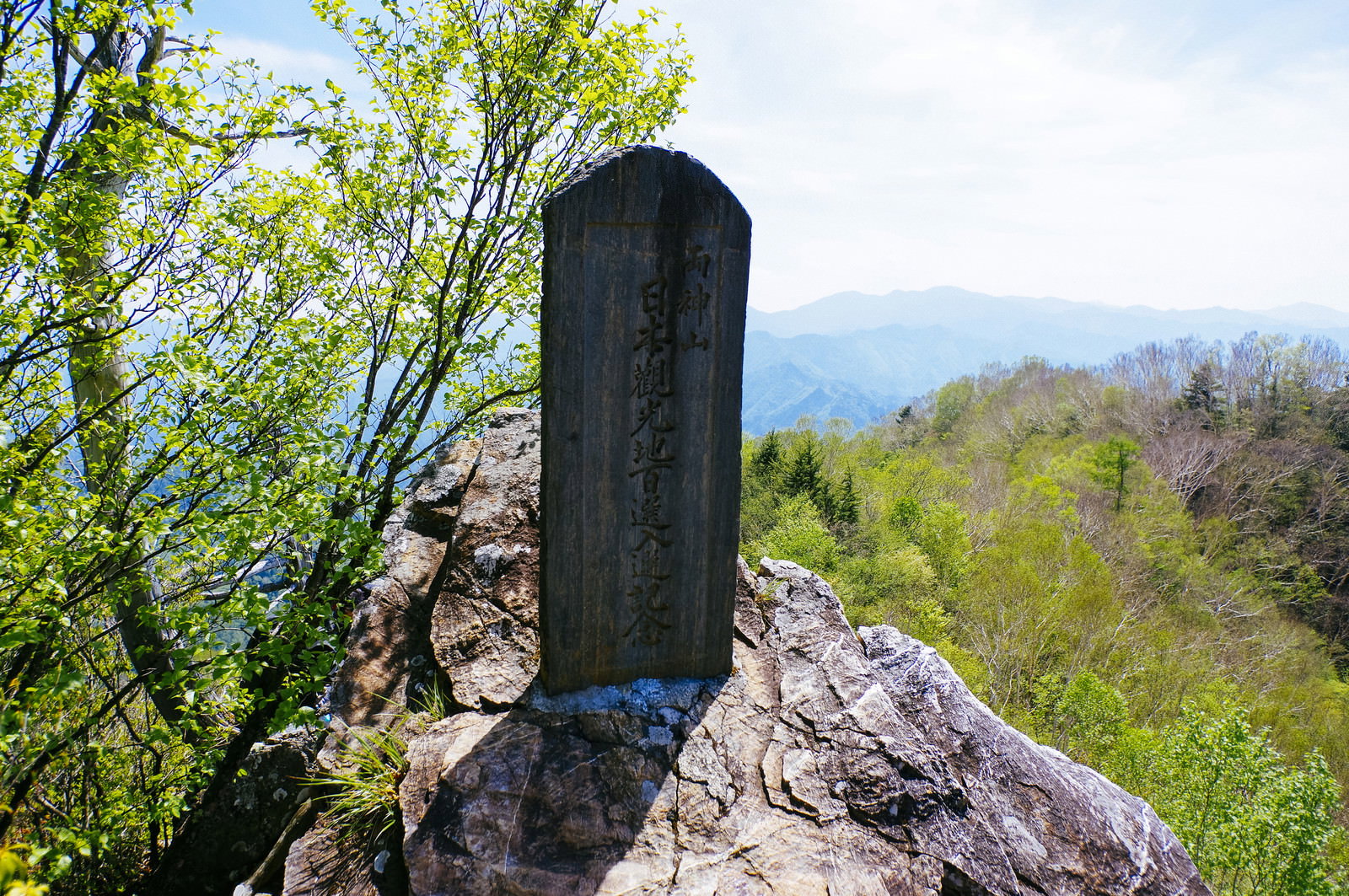 岩場の上に立つ両神山山頂の黒い石碑と新緑の山並み