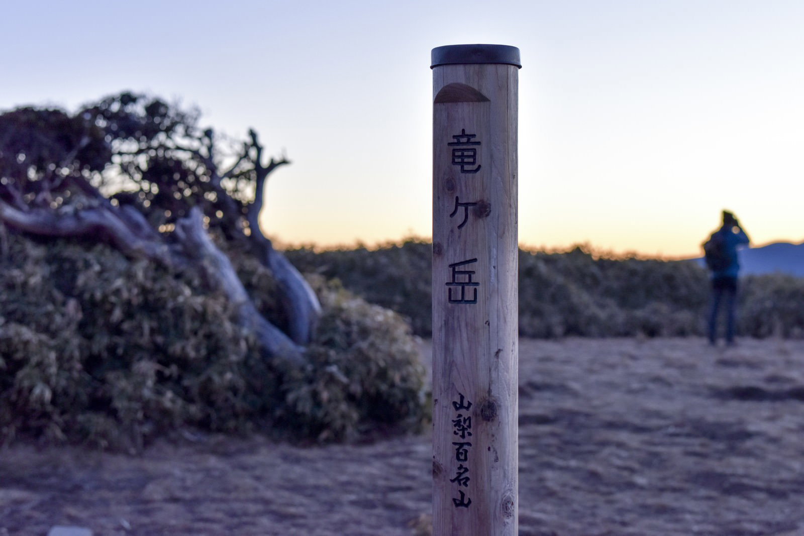 竜ヶ岳山頂の碑の手前に立つ標識と、朝焼けの中で山を眺める登山者