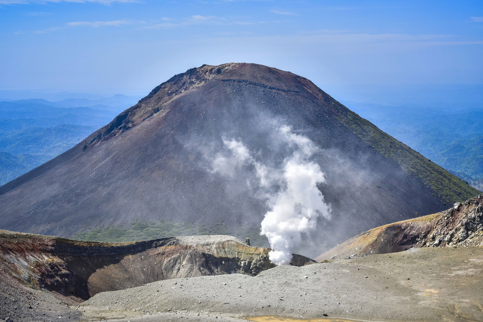 青空の下、火山地帯から立ち上る白い噴煙と背景の阿寒富士