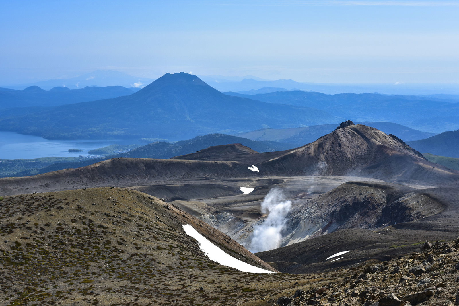 火山の複数の火口から白い蒸気が立ち上り、背景に雄阿寒岳を含む連山が青く霞んで見える風景
