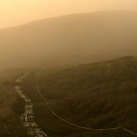 ガスと霧に包まれる夕暮れの登山道、オレンジ色の夕日が照らす山肌の写真