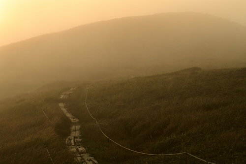 ガスと霧に包まれる夕暮れの登山道、オレンジ色の夕日が照らす山肌