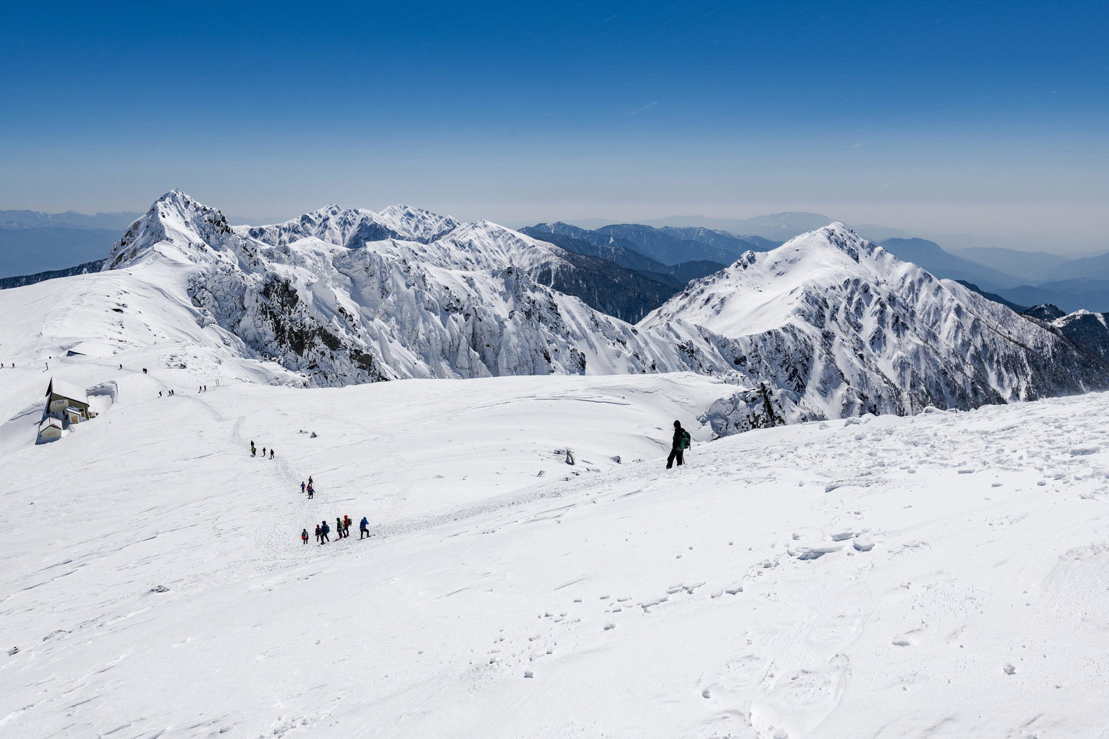 雪に覆われた中央アルプスの山々と青空の下を歩く登山者たち
