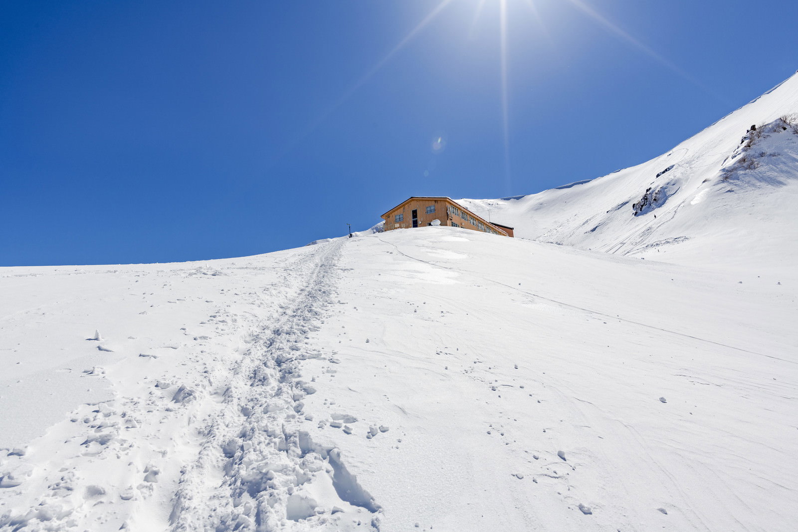 雪原の斜面に足跡が続き、青空の下に山小屋が見える冬山の風景