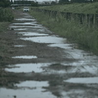 雨上がりの河川敷の砂利道に点在する水溜りと営業車の写真