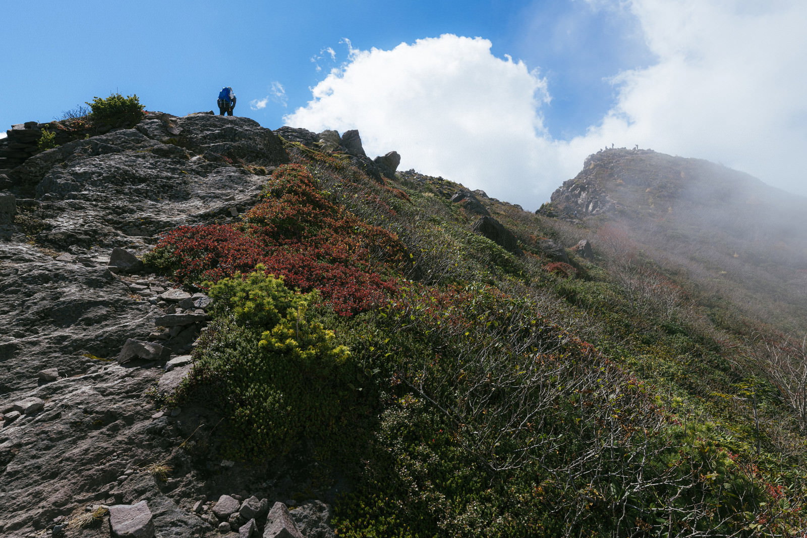 紅葉した外輪山の岩場を歩く登山者と青空に浮かぶ白い雲