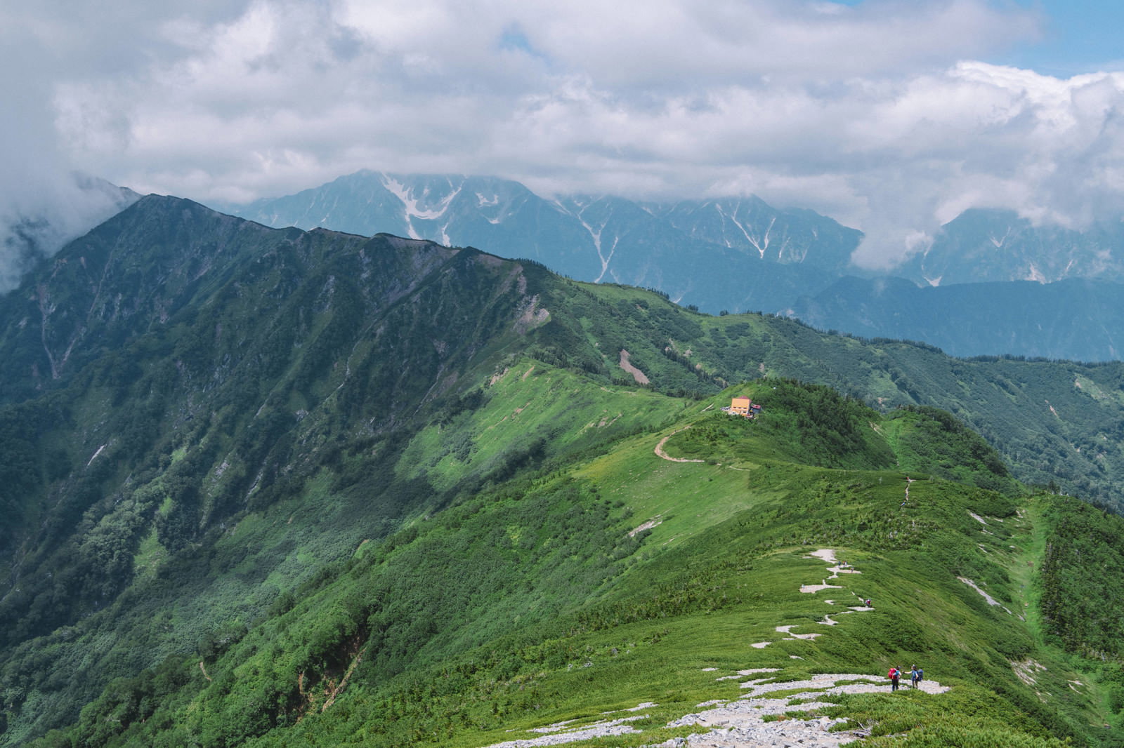 緑の稜線上に建つ種池山荘と背景に広がる立山連峰の山々