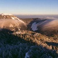 朝焼けに染まる冬の蓼科山、雪化粧した山頂を滝雲が流れるの写真