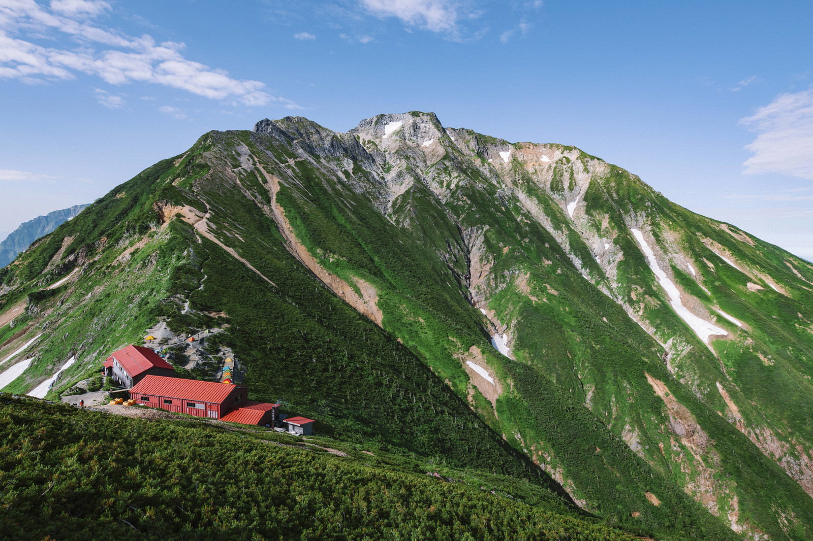 緑の山肌に白い雪渓が残る五竜岳と手前に建つ赤い屋根の五竜山荘