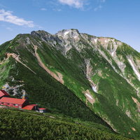 緑の山肌と白い雪渓が残る五竜岳と赤い屋根の五竜山荘の写真
