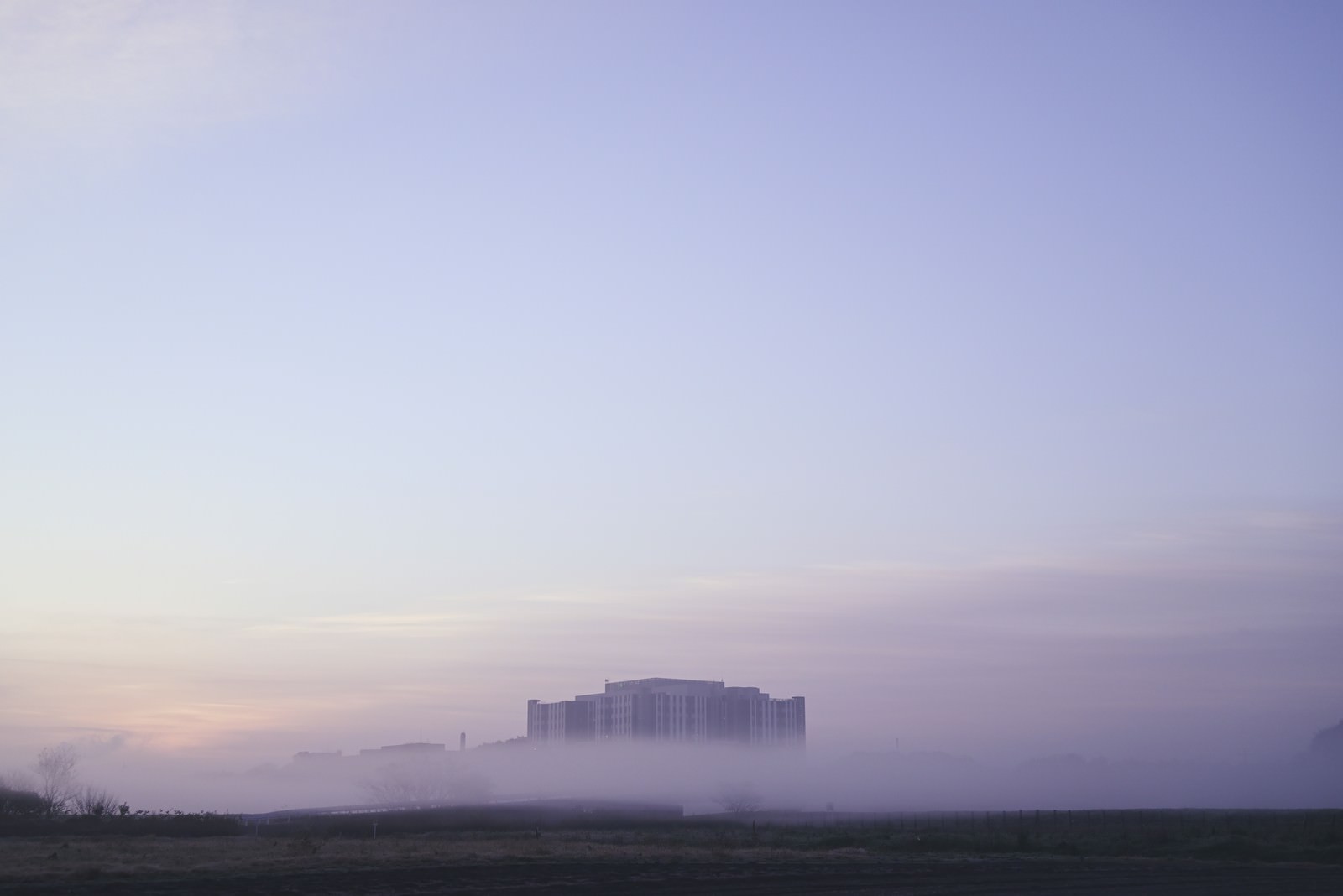朝霧に包まれた土手の向こうに建物が見える朝焼けの風景